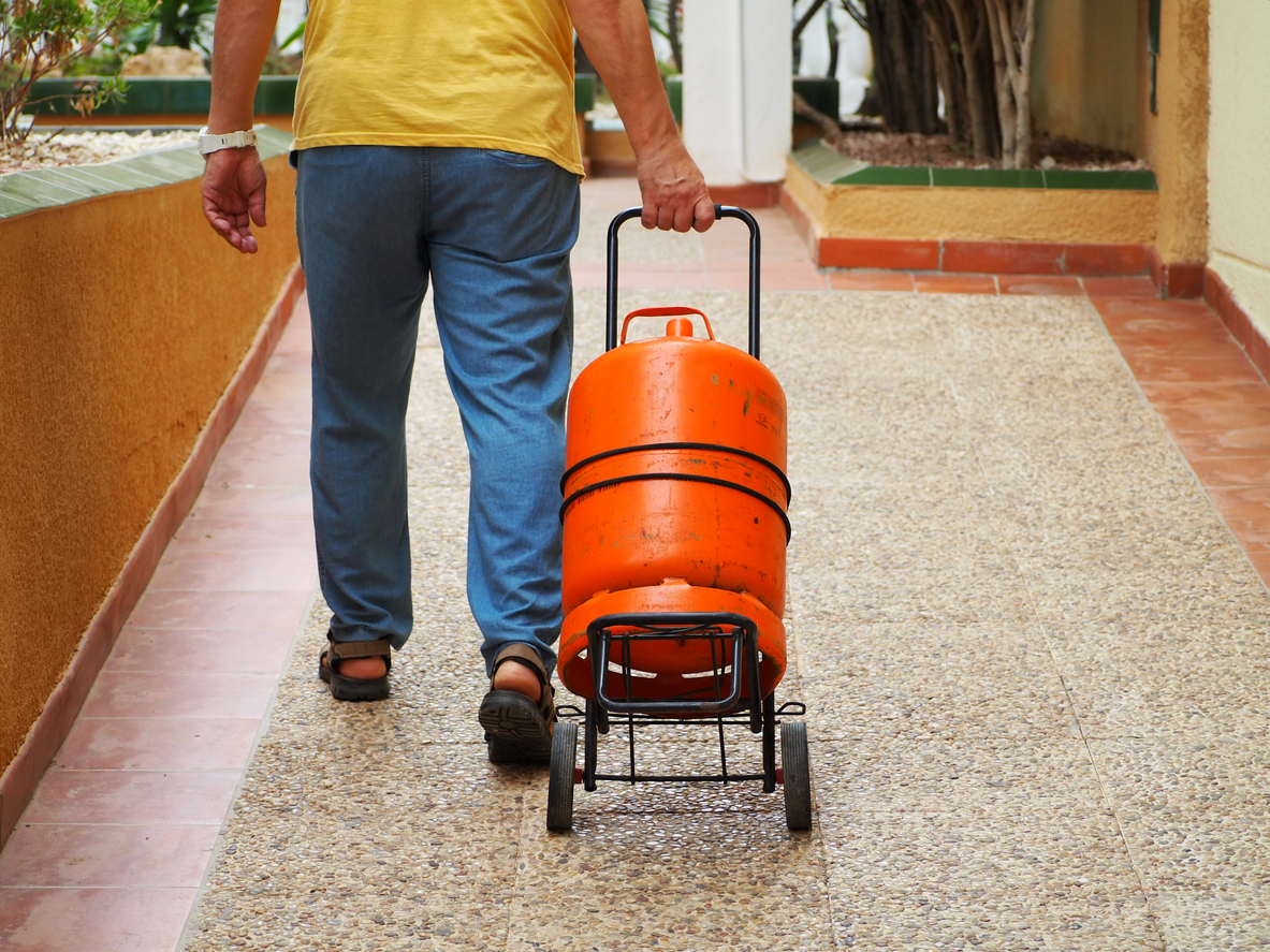 A man on a cart carries an orange gas cylinder along the street of a city.