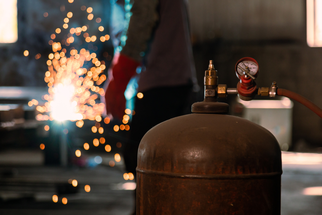 Closeup of Steel Oxygen Cylinder with industrial welder with a torch welding metal profiles in background